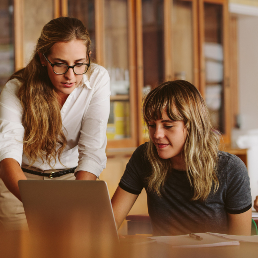 Two women are shown looking at a laptop on a desk.