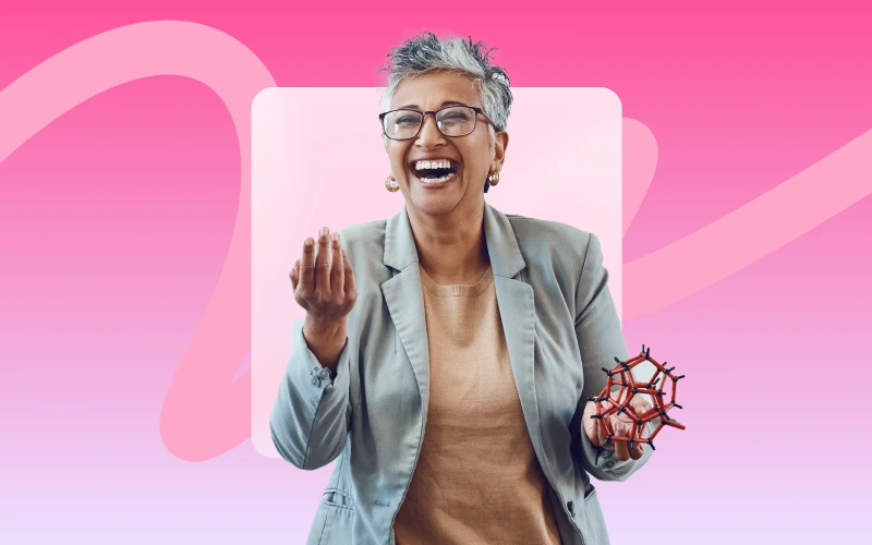 A gray-haired woman in glasses and a blazer laughs heartily while holding a red and black molecular model in her left hand.