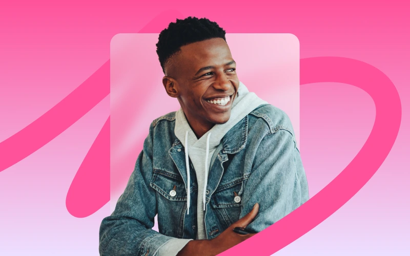 A young man with short dark hair and a wide smile looks off to his right, wearing a denim jacket over a light-colored hoodie, against a pink background.