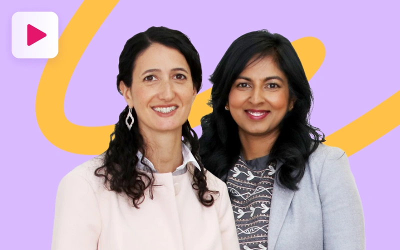 Two women, one with dark wavy hair and a light pink blazer, and the other with dark straight hair and a patterned top under a grey blazer, smile at the camera against a purple and orange background with a play button icon in the top left.