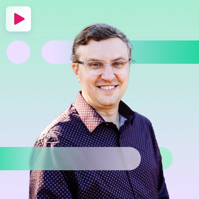 A smiling man wearing clear-rimmed glasses and a dark, patterned button-up shirt looks at the camera against a green, purple, and white abstract background with a play button icon in the top left.