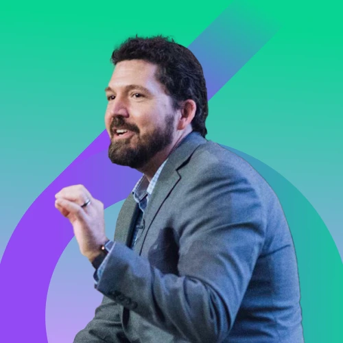 A bearded man in a grey suit jacket and blue collared shirt looks to his left with a slight smile and an open mouth, raising his left hand, against a green and purple abstract background.