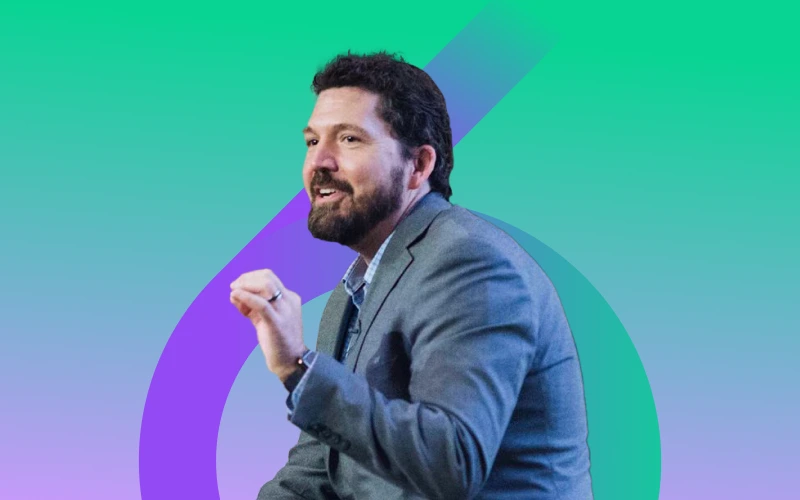 A bearded man in a grey suit jacket and blue collared shirt looks to his left with a slight smile and an open mouth, raising his left hand, against a green and purple abstract background.